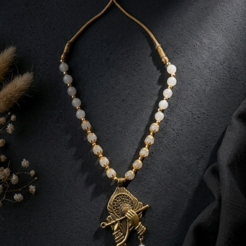 May include: A necklace featuring a series of smooth, round, off-white beads interspersed with small, ornate gold-toned metal spacers. The pendant is a detailed gold-toned metal design depicting a hand holding a flute against a circular frame.