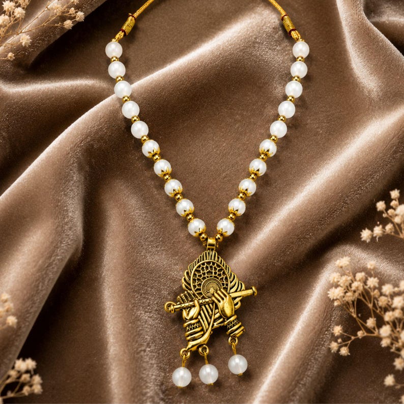 May include: A necklace featuring a string of white spherical beads and a gold-toned pendant depicting hands holding a flute. The pendant has an intricate circular design behind the hands and three small white beads hanging from the bottom edge.
