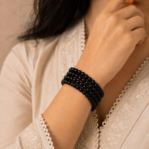 Puede incluir: Una pulsera de múltiples hilos compuesta por cuatro filas de cuentas esféricas negras, lisas y pulidas. Las cuentas son de tamaño uniforme y están ensartadas para crear una apariencia en capas alrededor de la muñeca, con un acabado brillante.