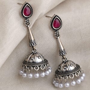 Puede incluir: Un par de pendientes jhumka de plata oxidada con un poste en forma de lágrima con una piedra roja oscura, un conector cónico y una base en forma de campana adornada con una hilera de pequeñas cuentas blancas colgando del borde inferior.