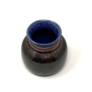 May include: A small, round ceramic vase with a dark brown glaze and a blue rim. The vase has a speckled texture.