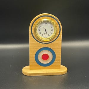 May include: A light-coloured wooden desk clock with a rounded top and a flat base. The clock face is white with black numerals and gold-tone hands, set in a gold-tone bezel. Below the clock is a circular target graphic in red, white, and blue.