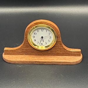 May include: A mantel clock featuring a light brown wooden frame with a curved top and a rectangular base. The clock face is white with black Arabic numerals and thin black hands, surrounded by a circular gold-toned metal bezel.