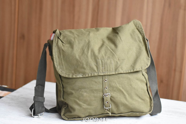 May include: A green canvas messenger bag with a black strap and a silver buckle. The bag has a flap with a single stitch detail.