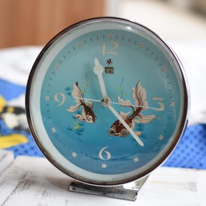 Op de afbeelding: Een zilveren wekker met een blauwe wijzerplaat en illustraties van gouden vissen. De wijzerplaat heeft zwarte cijfers en wijzers. De klok is ingesteld op 10:10.