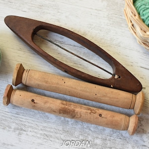 Könnte beinhalten: Zwei Holzspulen und eine Holzspindel zum Spinnen von Garn. Die Spulen sind hellbraun und haben eine dekorative Rille um die Ober- und Unterseite. Die Spindel ist dunkelbraun und hat einen Metallhaken. Im Hintergrund befindet sich eine Kugel grünes Garn.