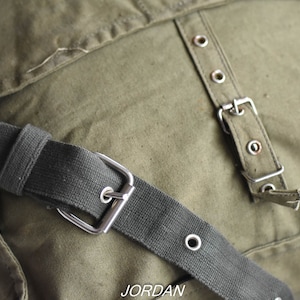 May include: A green canvas messenger bag with a black adjustable strap and silver buckle. The bag has a flap with a row of silver eyelets and a silver buckle.