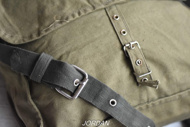 May include: A green canvas messenger bag with a black adjustable strap and silver buckle. The bag has a flap with a row of silver eyelets and a silver buckle.