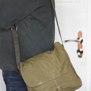 May include: A green canvas messenger bag with a long strap and metal buckles. The bag is worn over the shoulder of a person wearing a dark green sweater and blue jeans.
