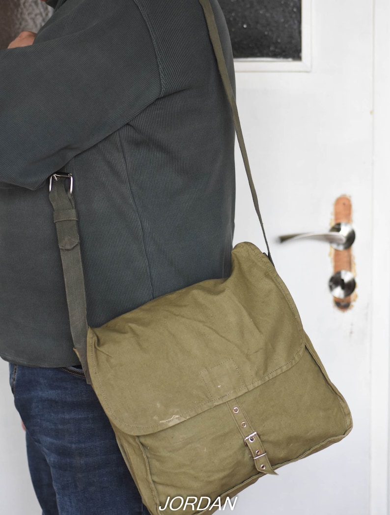 May include: A green canvas messenger bag with a long strap and metal buckles. The bag is worn over the shoulder of a person wearing a dark green sweater and blue jeans.