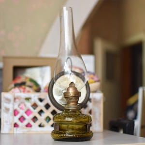 Puede incluir: Una lámpara de aceite vintage con una base de vidrio texturizado en color verde oliva y una chimenea de vidrio transparente y alta. La lámpara tiene un quemador metálico con una perilla de ajuste, situada frente a un fondo borroso.