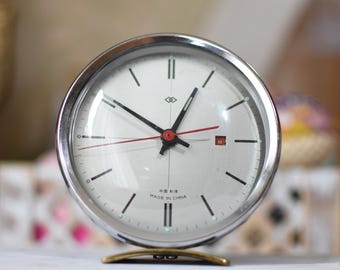 May include: A round analog table clock with a polished silver-tone metal frame and a white face. It features black hour markers, black hour and minute hands, a thin red second hand, and a small red date window. The text "MADE IN CHINA" is printed.