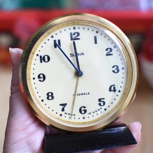 May include: A gold-colored analog clock with black hands and a white face. The clock face has black numbers and the words "SLAVA" and "17 JEWELS" printed on it. The clock is on a black stand and has the words "MADE IN USSR" printed on the bottom.