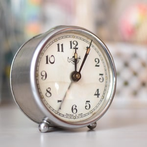 May include: A vintage silver-toned alarm clock with a cream-colored face. The clock features black numerals, hands, and a small logo at the top. The clock is set to approximately 1 o'clock. The clock is small, with a diameter of about 3 inches.