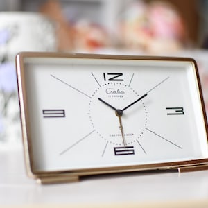May include: A rectangular, gold-coloured alarm clock with a white face. The clock has black numerals at 12, 3, 6, and 9 o'clock, with thin black lines radiating from the centre. The clock face has the brand name "Gaba" and "Copenhagen" printed on it.