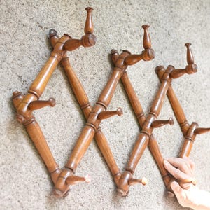 Puede incluir: Un perchero de pared extensible de madera con un diseño de celosía y múltiples ganchos para colgar objetos. El soporte está fabricado en madera de tono medio con un acabado liso y dispuesto en un patrón en zigzag ajustable en anchura.