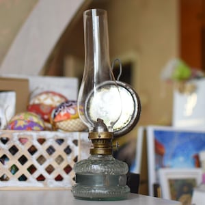 Puede incluir: Una lámpara de aceite vintage con una chimenea de vidrio transparente, un mecanismo de quemador de latón y una base de vidrio translúcido texturizado. La lámpara incluye un reflector de metal circular unido a la parte posterior del quemador.