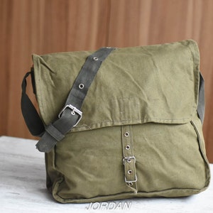 May include: A green canvas messenger bag with a black adjustable strap and silver buckles. The bag has a flap closure and a zippered pocket on the front.