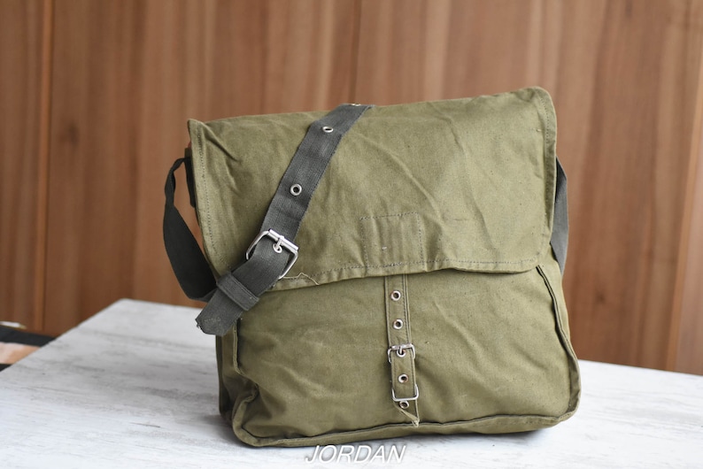 May include: A green canvas messenger bag with a black adjustable strap and silver buckles. The bag has a flap closure and a zippered pocket on the front.
