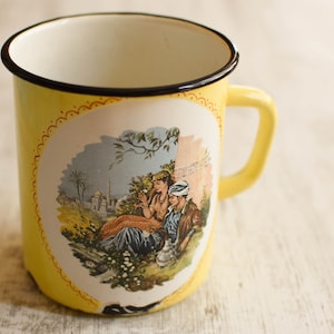 May include: Yellow enamel mug with a black rim and a colorful illustration of a couple in a landscape setting. The mug has a red and white decorative border.