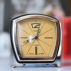 Può includere: Una sveglia vintage Slava con quadrante quadrato beige, indici neri e cornice in metallo argentato. Il quadrante riporta le scritte SLAVA, 11 JEWELS e MADE IN USSR, con lancette nere e una lancetta dei secondi di colore blu.