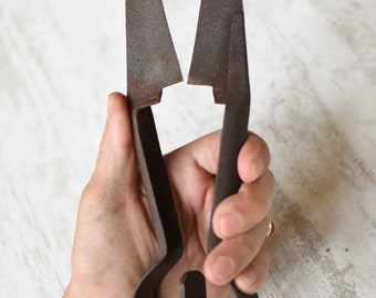 Könnte beinhalten: Antike Metallschere mit rostiger Oberfläche, gebogenen Griffen und dreieckigen Klingen. Die Klingen sind geöffnet und zeigen ihre scharfen Kanten. Die Schere wird in einer Hand vor einem weißen, strukturierten Hintergrund gehalten. Ein Schneidwerkzeug.