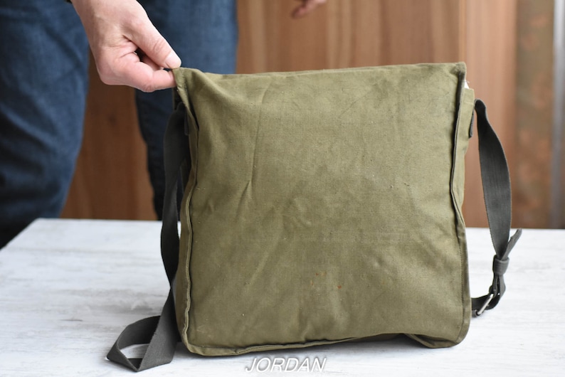 May include: A green canvas messenger bag with a black adjustable strap. The bag has a flap closure and a zippered pocket on the front.