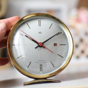 May include: A vintage, round alarm clock with a gold-colored bezel and a silver face. The clock has black hands, a red second hand, and a small date window. The clock is marked "Made in China".
