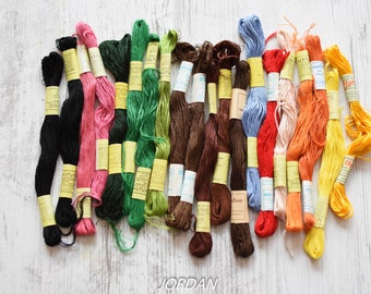 May include: Assortment of embroidery floss spools in various colors, including black, pink, green, brown, blue, red, orange, and yellow. The spools are arranged in a row on a white wooden surface.