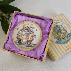Op de afbeelding: Een witte keramische herinneringsdoos met een geel deksel met een cartoonillustratie van dieren op een boot. Het deksel heeft de tekst "First Curl Keepsake Box".
