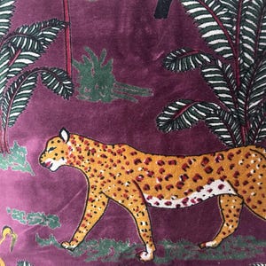 Op de afbeelding: Close-up van een bordeauxrode stof met een jungledesign. Het ontwerp toont een cheetah met oranje vlekken, toekans, apen en palmbladeren. De stof heeft een zachte, fluweelachtige textuur.