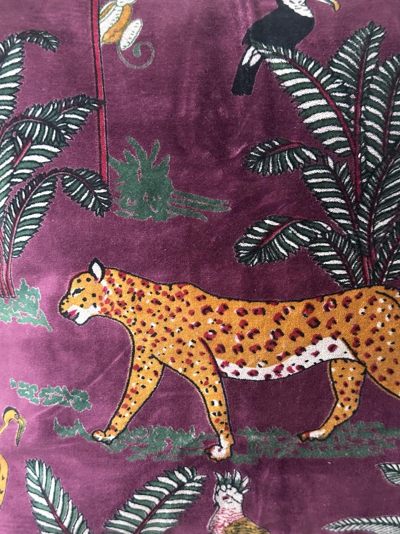 Op de afbeelding: Close-up van een bordeauxrode stof met een jungledesign. Het ontwerp toont een cheetah met oranje vlekken, toekans, apen en palmbladeren. De stof heeft een zachte, fluweelachtige textuur.