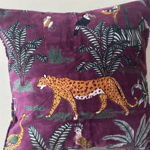 Op de afbeelding: Vierkantig pluche kussen met een dieppaarse achtergrond en een jungle-thema patroon. Het ontwerp omvat een lopende luipaard, toekans, een zebra en weelderig groen gebladerte. Het kussen heeft een zachte, fluweelzachte textuur.