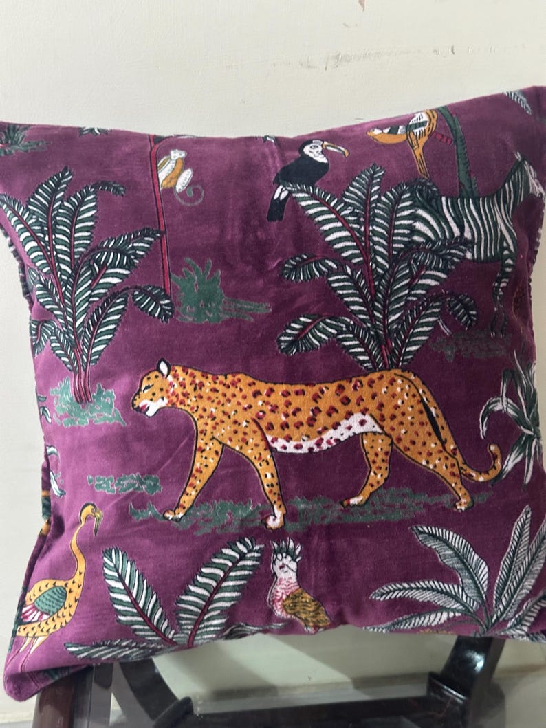 Op de afbeelding: Vierkantig pluche kussen met een dieppaarse achtergrond en een jungle-thema patroon. Het ontwerp omvat een lopende luipaard, toekans, een zebra en weelderig groen gebladerte. Het kussen heeft een zachte, fluweelzachte textuur.