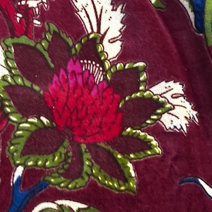 Puede incluir: Primer plano de una tela burdeos con un estampado floral. El diseño presenta una flor grande y estilizada en tonos rojos, rosas y verdes, con acentos de hojas blancas. La tela tiene una textura suave y aterciopelada, lo que sugiere un textil lujoso.