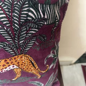 Op de afbeelding: Een decoratief kussen met een bordeauxrode achtergrond met een junglesc&egrave;ne. Het ontwerp omvat een cheetah met oranje vlekken, een zebra met zwart-witte strepen en groen-wit gebladerte.