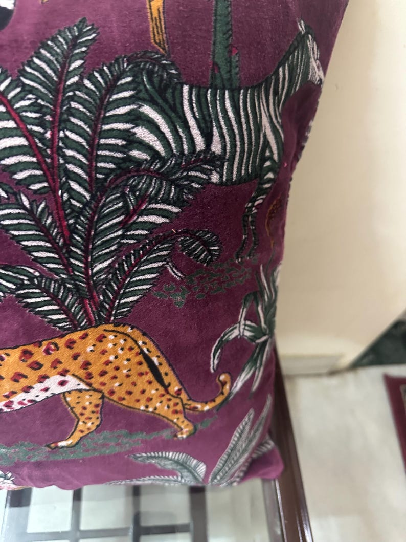 Op de afbeelding: Een decoratief kussen met een bordeauxrode achtergrond met een junglesc&egrave;ne. Het ontwerp omvat een cheetah met oranje vlekken, een zebra met zwart-witte strepen en groen-wit gebladerte.