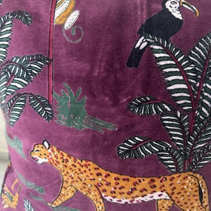 Op de afbeelding: Bordeaux fluwelen kussen met een junglepatroon. Toont een luipaard, een toekan en een aap te midden van groene bladeren. De luipaard is bruin met bruine vlekken. De toekan is zwart met een witte borst en gele snavel. De aap is wit en geel.