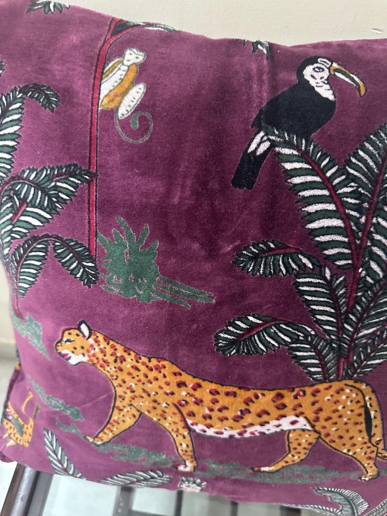 Op de afbeelding: Bordeaux fluwelen kussen met een junglepatroon. Toont een luipaard, een toekan en een aap te midden van groene bladeren. De luipaard is bruin met bruine vlekken. De toekan is zwart met een witte borst en gele snavel. De aap is wit en geel.