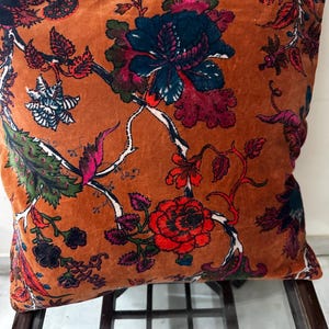 Op de afbeelding: Een bruin fluwelen kussen met een bloemenpatroon in tinten roze, rood, blauw en groen. Het kussen is versierd met een herhalend patroon van bloemen en bladeren.