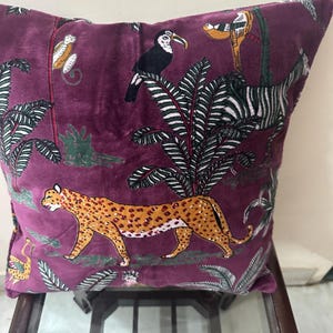 Op de afbeelding: Vierkantig sierkussen in een diepe paarse tint, met een jungle-thema ontwerp. Het ontwerp omvat een luipaard, zebra, toekan en aap te midden van weelderig gebladerte. Het kussen is waarschijnlijk gemaakt van fluweel of een vergelijkbare zachte stof.