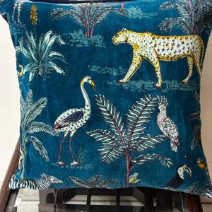 May include: Teal velvet cushion with a jungle design featuring a leopard, birds, and palm trees. The cushion has a dark blue velvet backing. The design includes a flamingo, toucan, and zebra.