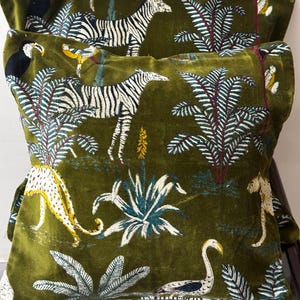 Könnte beinhalten: Zwei olivgrüne Samtkissen mit Dschungeltierdruck. Die Kissen zeigen Zebras, Leoparden, Tukane und andere Vögel inmitten üppigen Blattwerks. Das Design umfasst weiße, schwarze, gelbe und türkisfarbene Akzente.