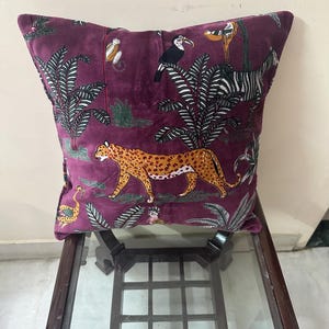 Op de afbeelding: Een vierkant, pruimkleurig sierkussen met een junglethema. Het kussen heeft een luipaard, toekan, zebra en aap te midden van weelderig gebladerte. Het kussen ligt op een donkere houten tafel met een glazen blad.