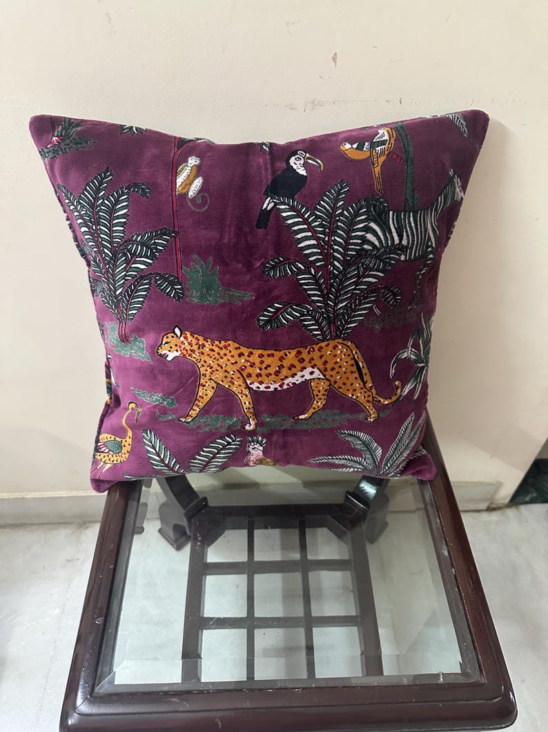 Op de afbeelding: Een vierkant, pruimkleurig sierkussen met een junglethema. Het kussen heeft een luipaard, toekan, zebra en aap te midden van weelderig gebladerte. Het kussen ligt op een donkere houten tafel met een glazen blad.