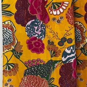 Könnte beinhalten: Nahaufnahme eines leuchtend gelben Stoffes mit einem kräftigen Blumenmuster. Das Design zeigt große, stilisierte Blumen in Fuchsia-, Burgunder-, Braun- und Türkistönen auf gelbem Hintergrund. Grüne Blätter und kleinere florale Akzente vervollständigen das Design.