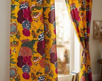 Op de afbeelding: Een paar kamerhoge gordijnen met een levendige mosterdgele achtergrond en een herhalend patroon van grote, gestileerde bloemen in tinten diep magenta, donkerblauwgroen en gedempt oranje, opgehangen aan een donkerbruine houten gordijnroede.