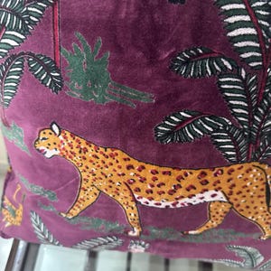 Op de afbeelding: Een decoratief kussen met een bordeauxrode achtergrond met een luipaardontwerp met oranje vlekken. Het kussen heeft ook groene en witte palmboom- en bladerpatronen. Het kussen is gemaakt van een zacht, fluwelig materiaal.