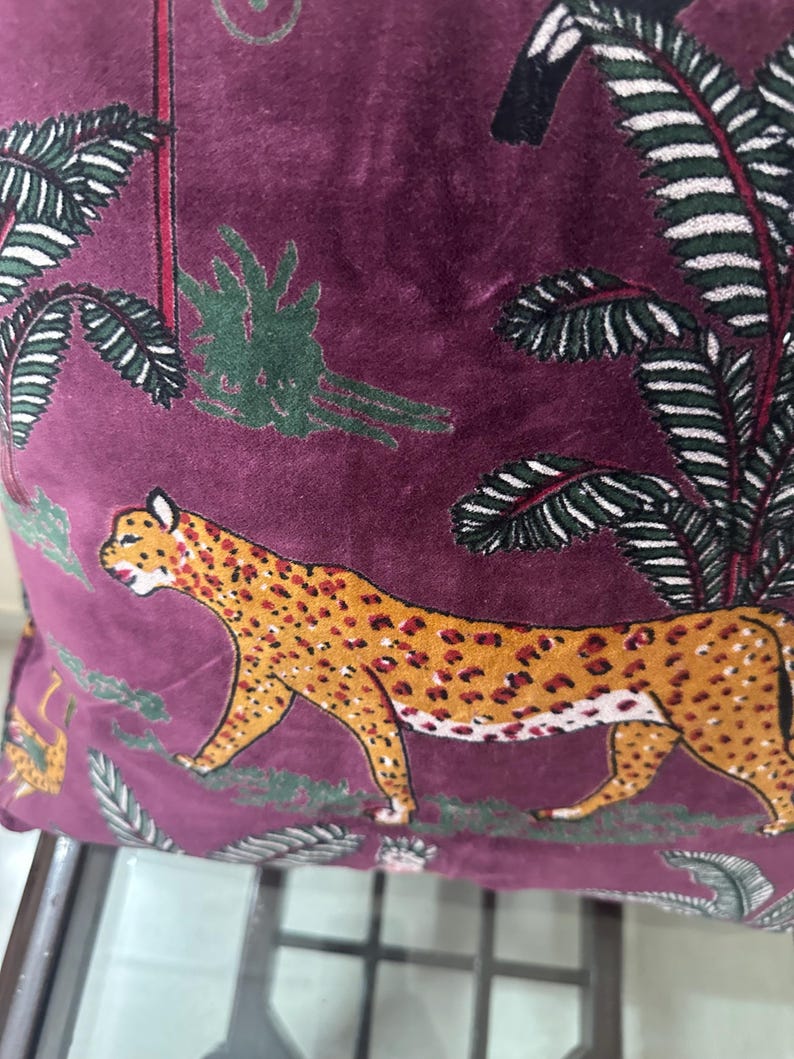 Op de afbeelding: Een decoratief kussen met een bordeauxrode achtergrond met een luipaardontwerp met oranje vlekken. Het kussen heeft ook groene en witte palmboom- en bladerpatronen. Het kussen is gemaakt van een zacht, fluwelig materiaal.