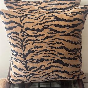 Può includere: Due cuscini quadrati con una base color beige e un motivo a strisce di tigre nere. Il tessuto ha un aspetto morbido e strutturato, e i cuscini sono impilati uno sopra l'altro, mostrando una stampa animalier coerente.